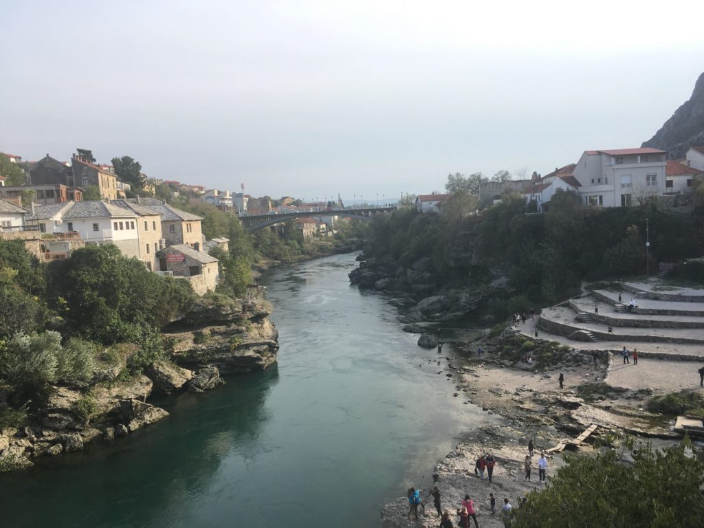 A beautiful landscape view from Mostar's Stari Most bridge, depicting a central winding river with local housing either side, and a simple bridge in the distance