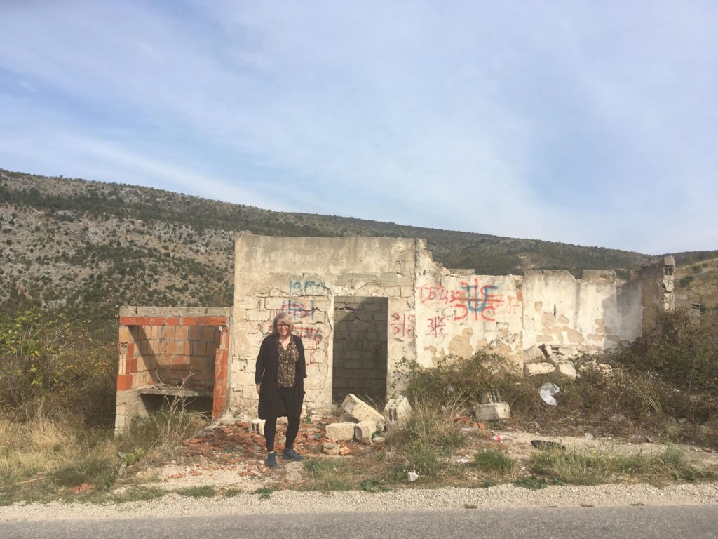 Photo of me stood in front of a small derelict brick building that was at the side of the road, that we pee'd in risking treading on landmines. It has no roof & graffiti in Bosnian on it. The Dalmatian mountains in the background.