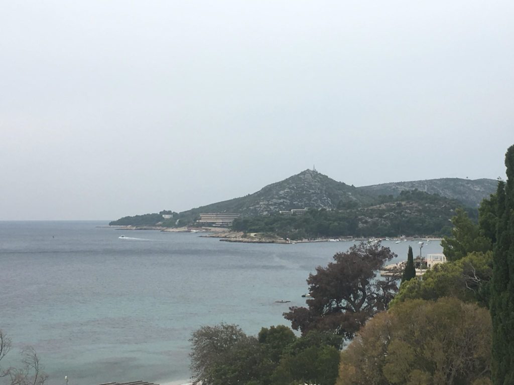 The Croatian dappled grey Dalmatian mountains in the far horizon, and a flat calm bay with a blue/grey sea. Trees in the foreground. A small speed boat in the water